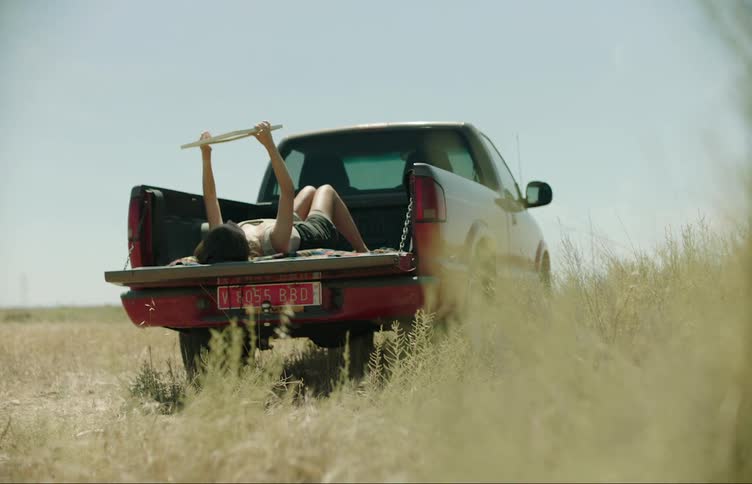 A woman lying in the back of a red pickup truck parked in a grassy area and she is holding a mirror and looking at herself in it