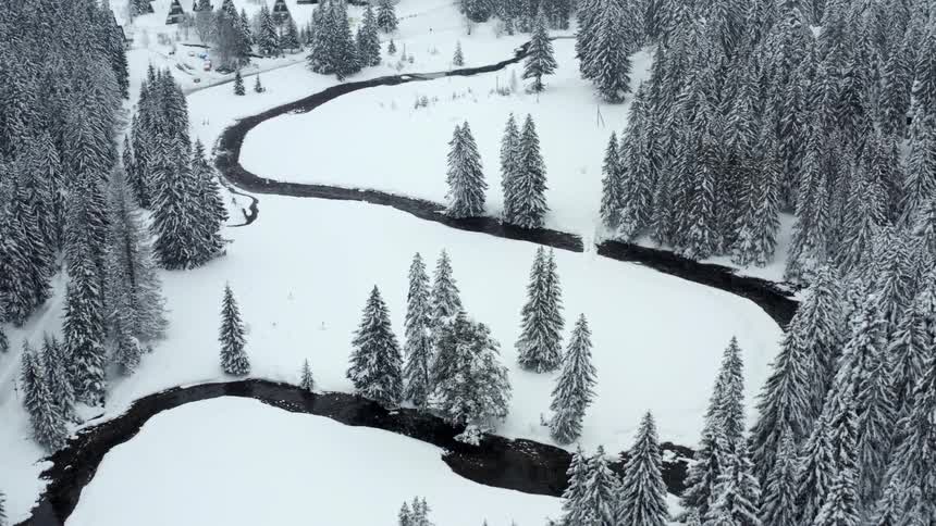 An aerial view shows a winding river flowing through a snowy winter landscape with dense coniferous forests