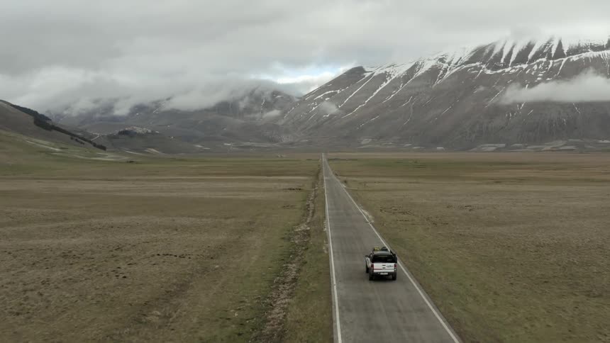 Vehicle drives along a road passing through grassland and towards snowy mountains on an overcast day