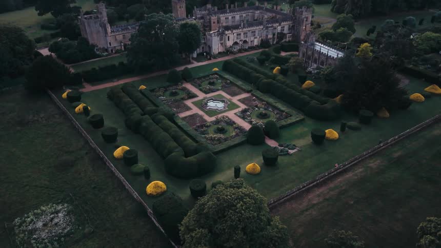 An aerial view shows a historic castle with manicured gardens and lush surrounding greenery