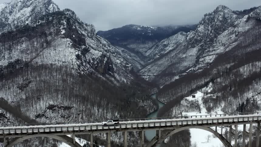 Lone truck crosses a narrow bridge standing tall over the river passing through a valley between mountains on a cloudy winter day