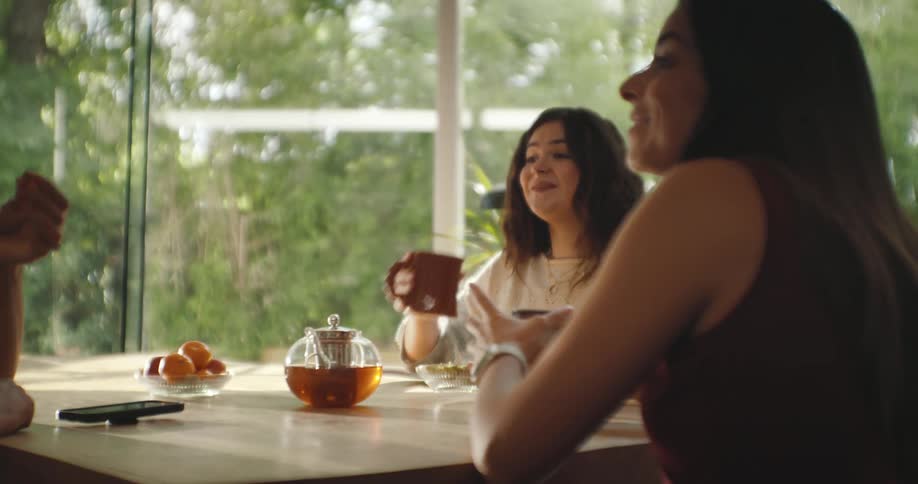 A group of three women gathered around a table and they are talking and one of them is drinking something from a mug