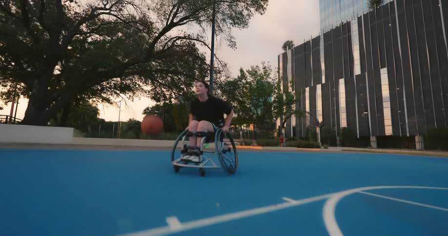 A person in a wheelchair dribbles a basketball on a blue court in front of a tall building