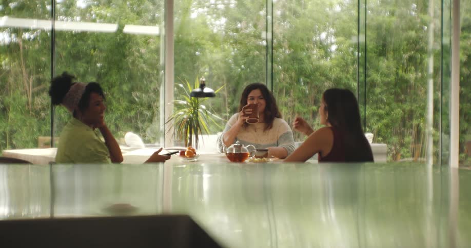 Three women sitting around a table drinking tea and having conversation as sunlight shines through the window