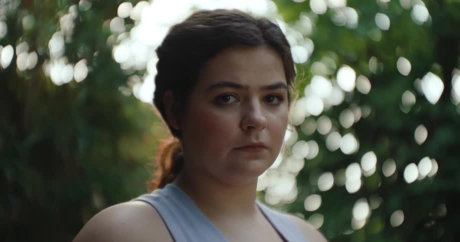 The face of a young woman who is standing and staring straight ahead and she is wearing a gray tank top