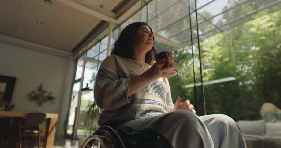 A young woman is wearing a coat and is sitting in a wheelchair while drinking something from a mug