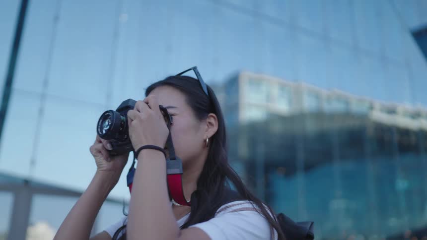 A woman with long dark hair takes a photo with a camera in front of a large glass building