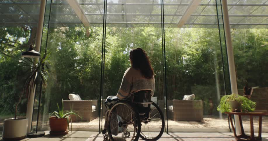 A woman sitting in a wheelchair in a living room in front of large glass windows