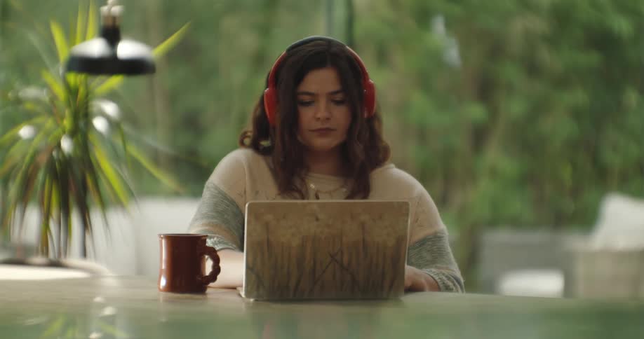A young woman sitting at a table using her laptop wearing headphones with a mug next to her