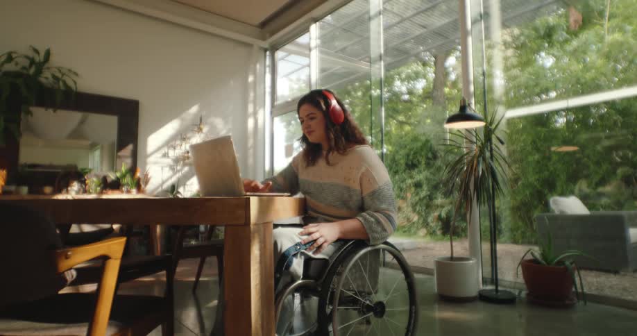 A woman in a wheelchair types on a laptop and listens to headphones while sitting at a table in a house