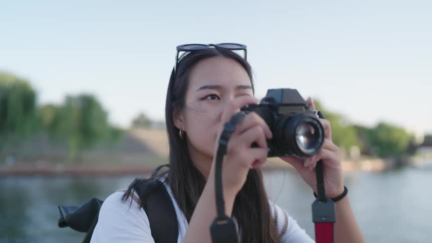 A woman standing with her back to a body of water in a city park holds up her camera and takes a photo