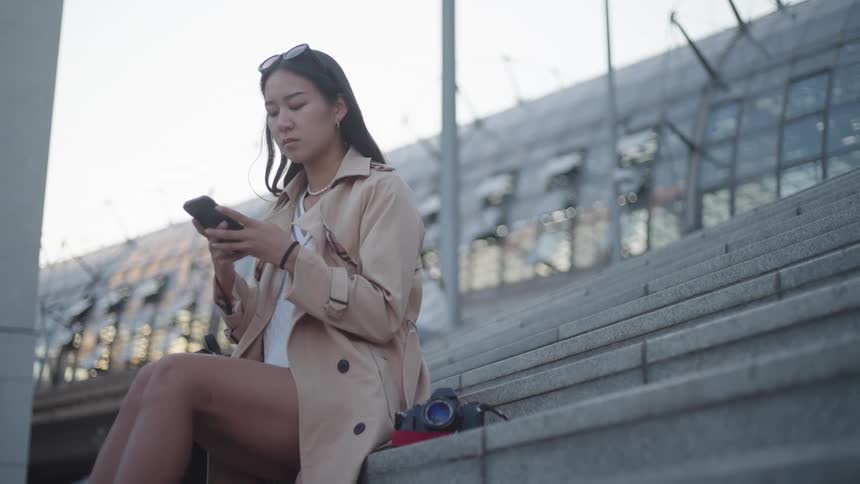 A woman sitting on concrete steps looking at her smart phone then looking up