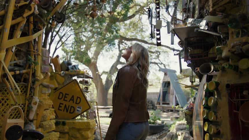 A young woman walks through a narrow alley filled with cluttered objects in a natural setting