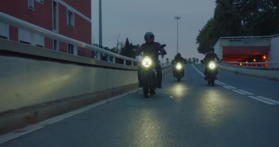 Three motorcyclists ride down a city street at dusk