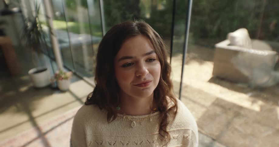 A smiling female teenager sits inside a house passing a basketball from one hand to another