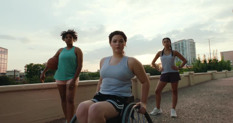 Two women standing around a third woman sitting in a wheelchair outdoors on a sidewalk
