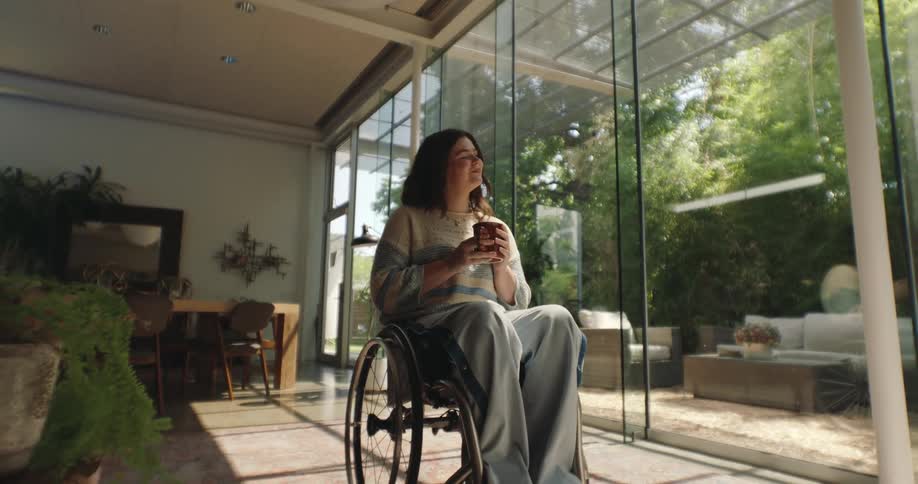 A woman in a wheelchair holds a cup inside a room with large glass windows
