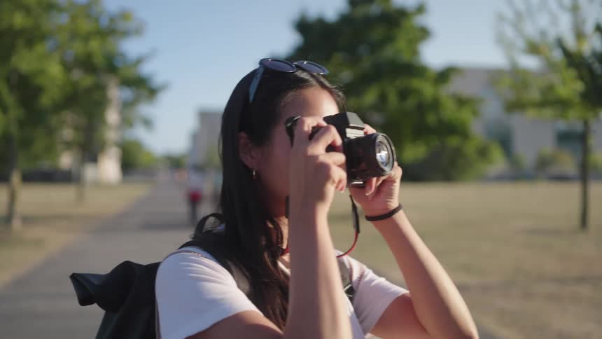 A woman walking in the park stops and pulls up a camera to take a photo then keeps walking in the direction in which she pointed the camera