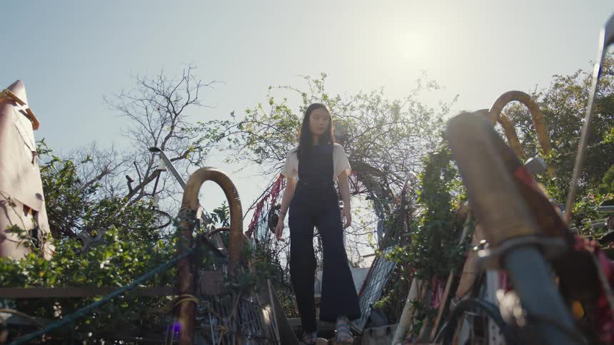 A woman stands at the top of an outdoor staircase surrounded by lush vegetation and an array of metallic pipes