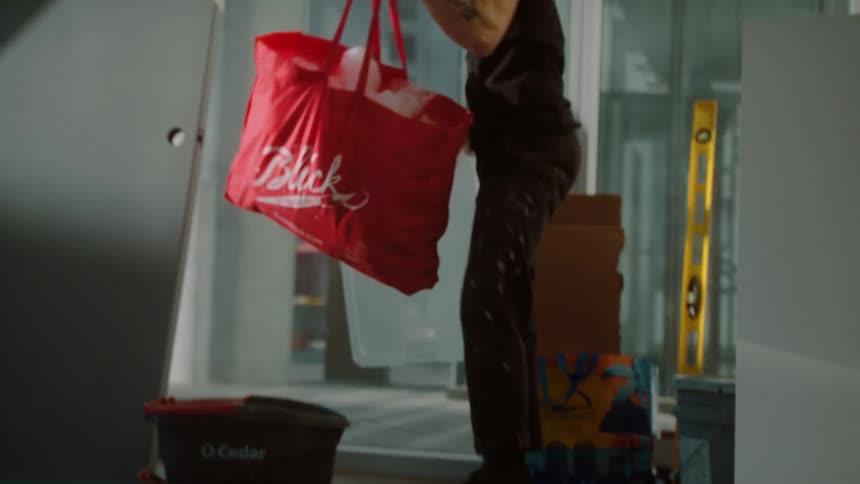 A man walking and bending over to pick up a red bag off the floor in a bright sunny room