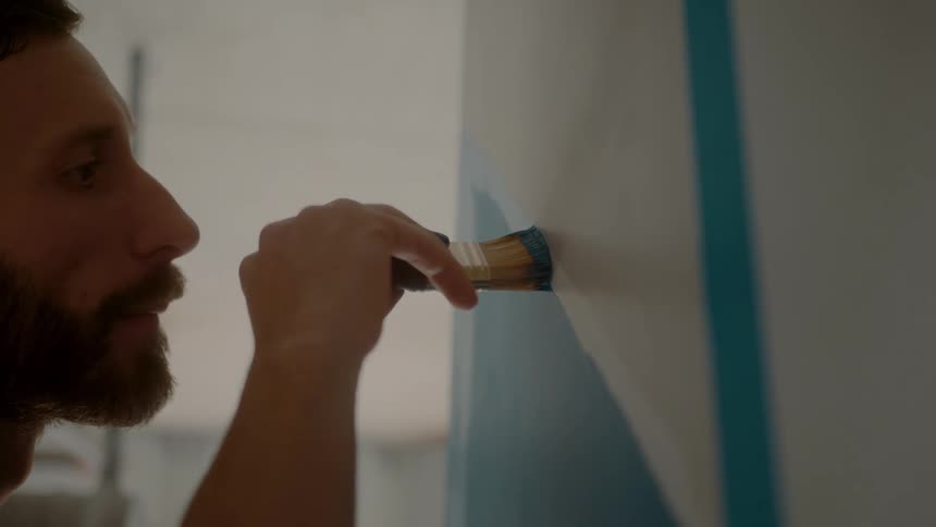 A bearded man painting a white wall with a brush with blue paint