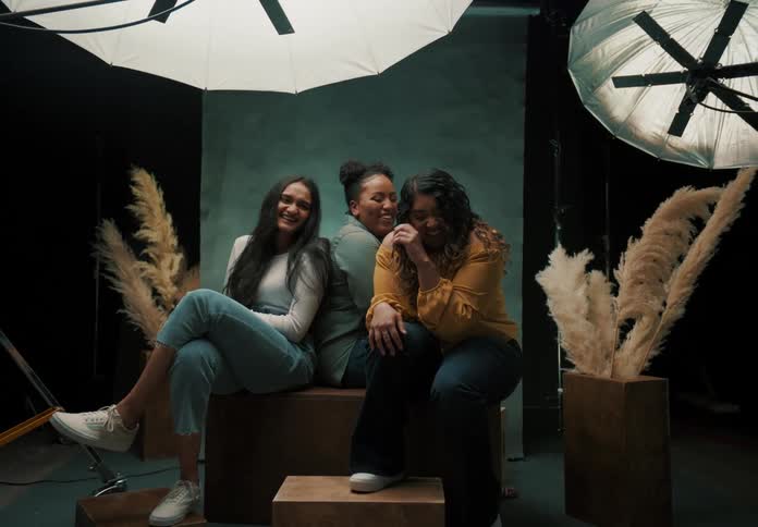 Three female friends are sitting on a studio bench with lights shining on them and they are happy and laughing