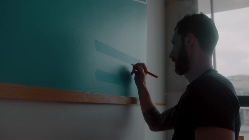 A man standing in front of a wall writing on a blue board as daylight shines through the windows