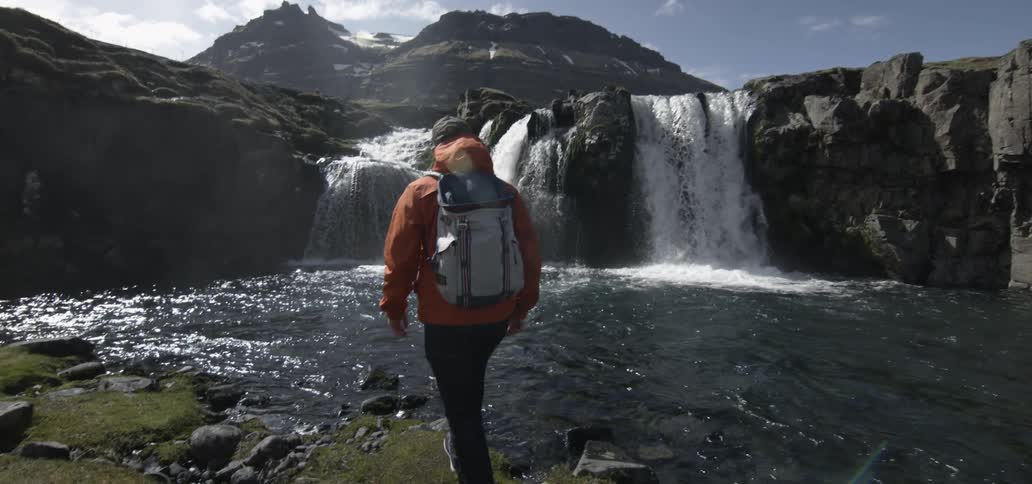 A hiker approaches a mountain lake and waterfalls