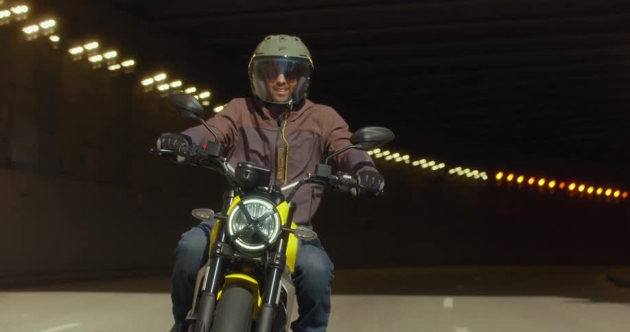 A man with a big smile rides a motorcycle through a tunnel and then down the street