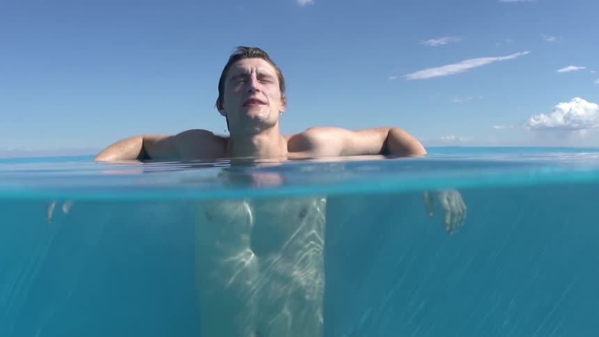 A young shirtless man has his body partially in the water of a swimming pool