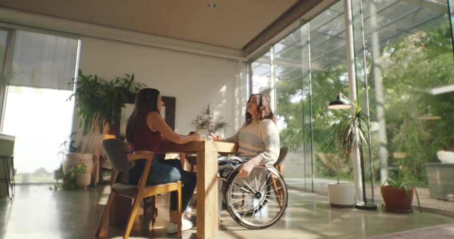 Two women sitting at a table conversing in a large space surrounded by large windows letting the sunlight in