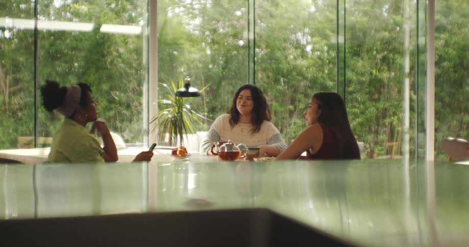 Three women sit at a dining table drinking tea and converse with each other