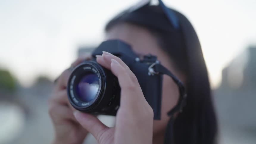 A woman is holding a camera lens up to their face with an urban background softly out of focus