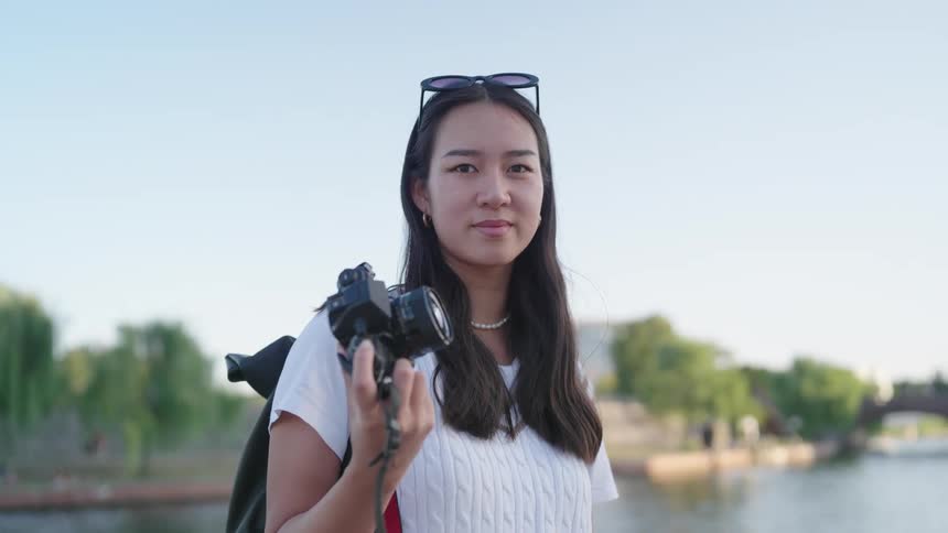 A young woman with sunglasses hanging in her hair looking forward and she is holding a digital camera