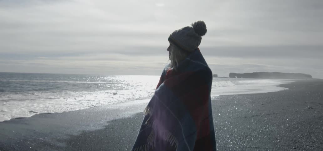 A woman that is wrapped in blanket is walking on the beach
