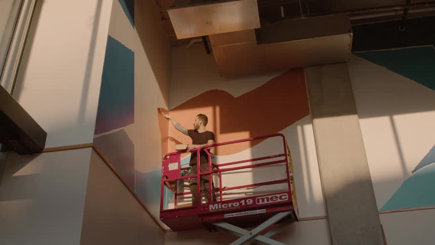 A worker on a lift paints graphics on the interior wall of a building