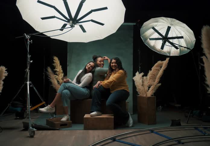 Three women sitting on a bench and are happy and looking straight ahead and at a backdrop around them with studio lights