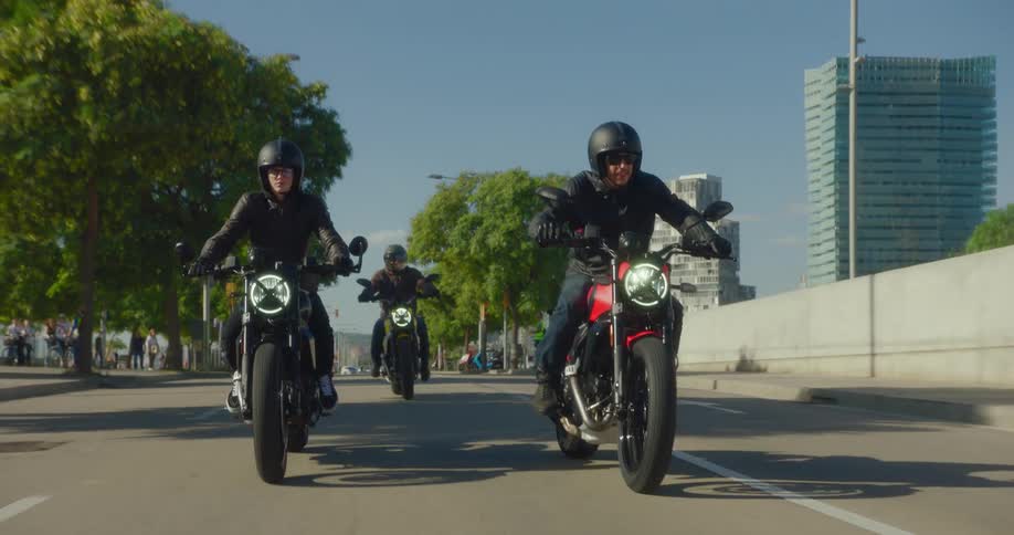 A group of motorcyclists ride down a city street lined with trees and buildings