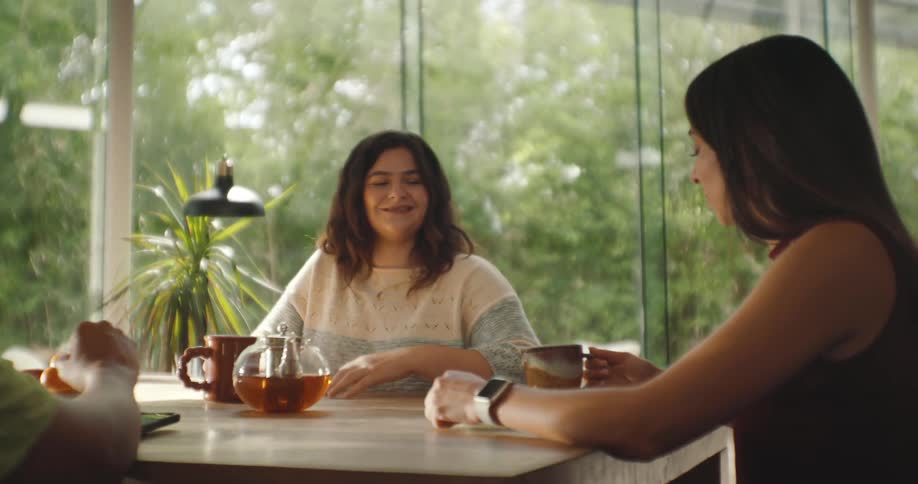 A group of three women who are gathered around a table and one of them is pouring a liquid into a mug and they toast