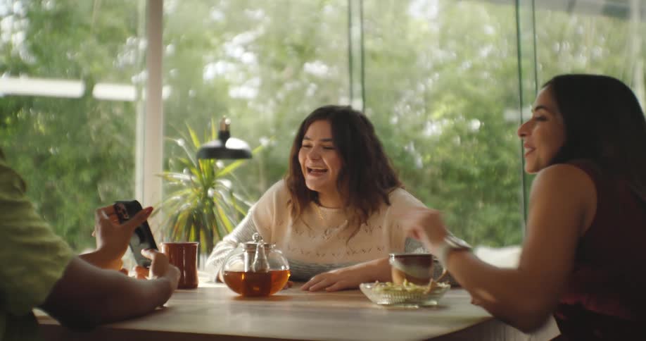 A group of three women around a table with breakfast foods and they are laughing while talking and one of them shows something from a cell phone and they laugh again