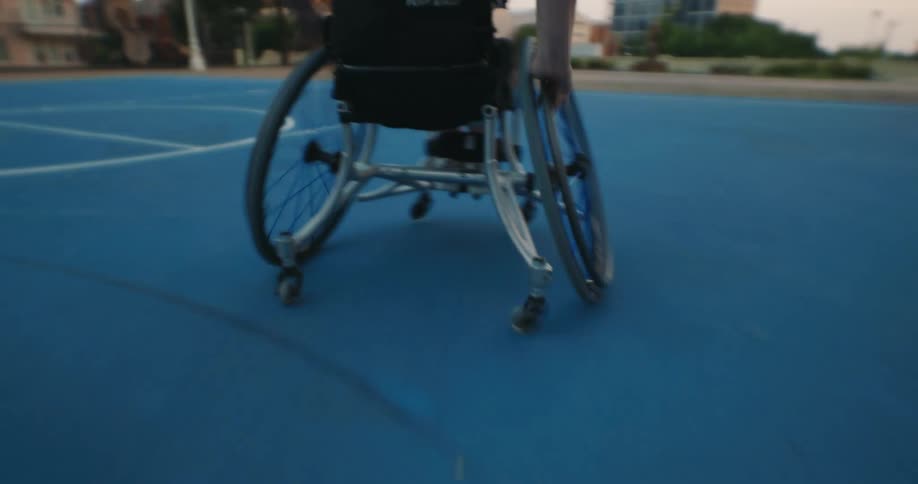 A woman in a wheelchair dribbles a basketball as she moves around a blue outdoor court at sunset goes for a shot but misses