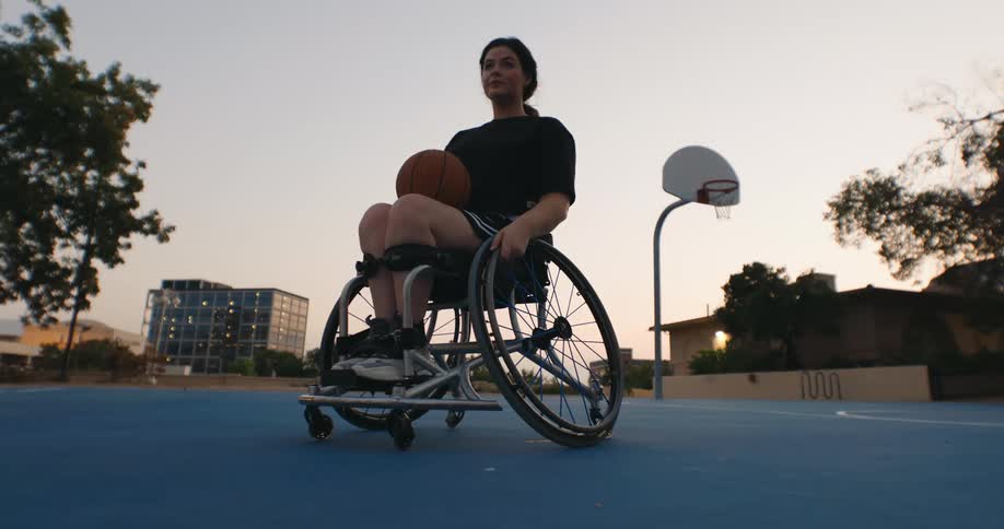 A woman in a wheelchair holds a basketball while sitting on a blue outdoor court at sunset with a basketball hoop in the background and trees and buildings nearby