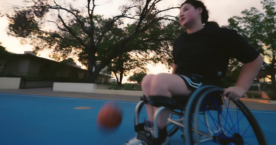 A young woman in a wheelchair dribbles and bounces a basketball around on a court