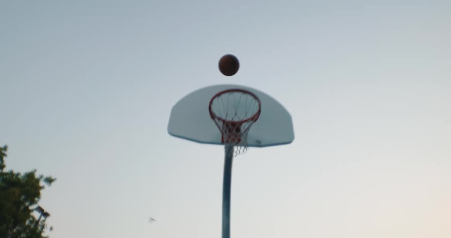 A man in a wheelchair moving down a basketball court outside at dusk as he shoots the ball