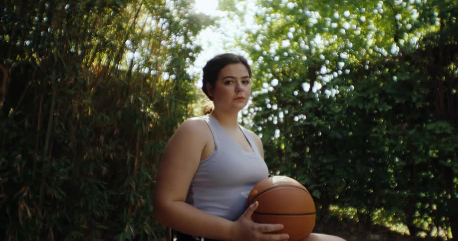 A woman sitting in a wheelchair holding a basketball in front of dense green foliage in a garden or park