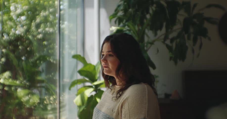 A woman is standing indoors next to a large window with sunlight streaming in surrounded by green plants