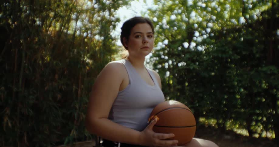 A teenage girl sits in a wheelchair while holding a basketball in a garden