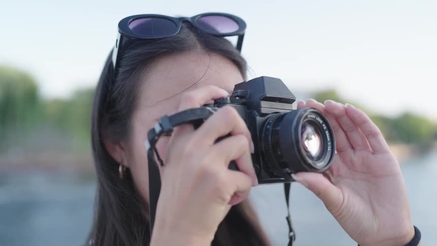 A young woman with glasses hanging from her hair is holding a black camera taking pictures and adjusting the lens with her finger