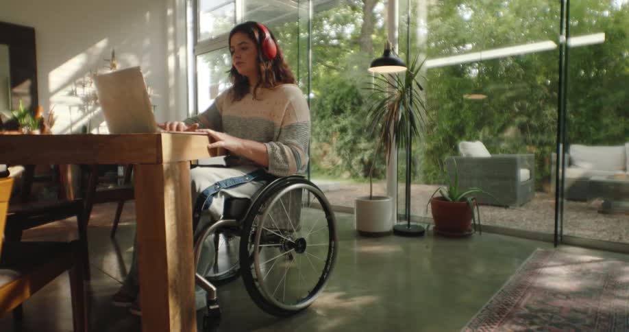 A young woman with headphones is in a wheelchair using a laptop in a room interior