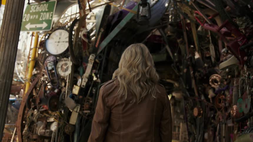 A blonde woman from behind wearing a leather jacket is entering a place full of junk on the sides